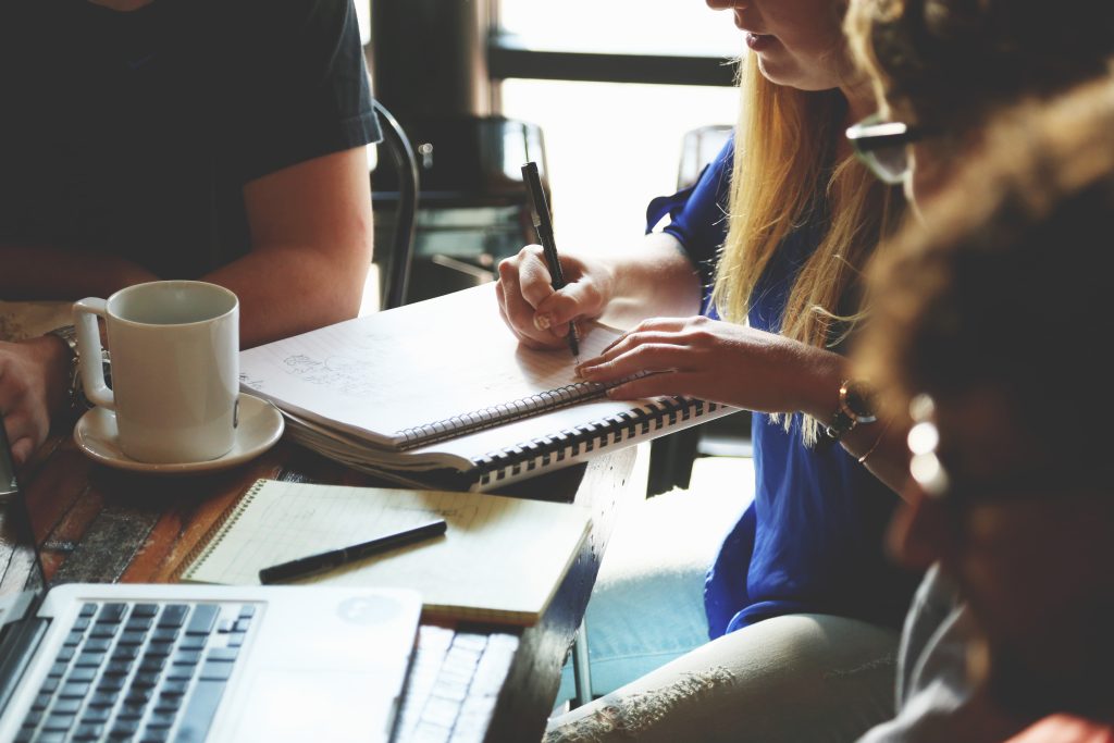 A group brainstorming session in a cafe, with a woman writing notes in a notebook, coffee cup, and laptop visible on the table.