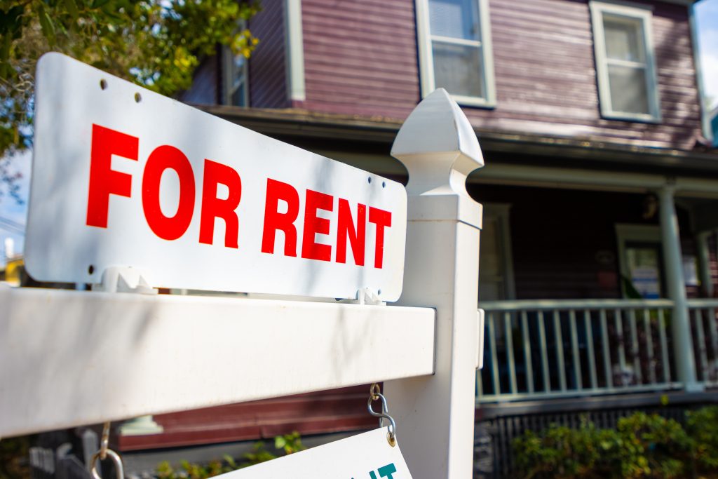 A "For Rent" sign prominently displayed in front of a large, purple house, indicating available rental property.