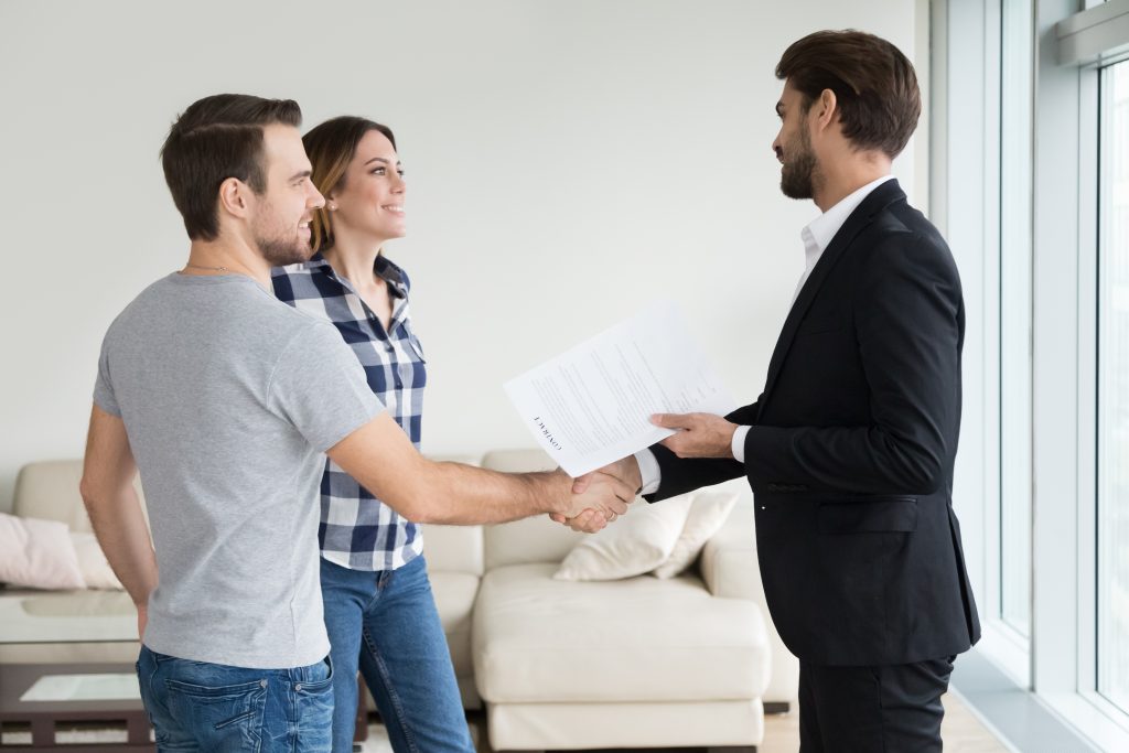 A man in a suit shakes hands with a couple in a modern living room while holding a document, suggesting a business agreement.